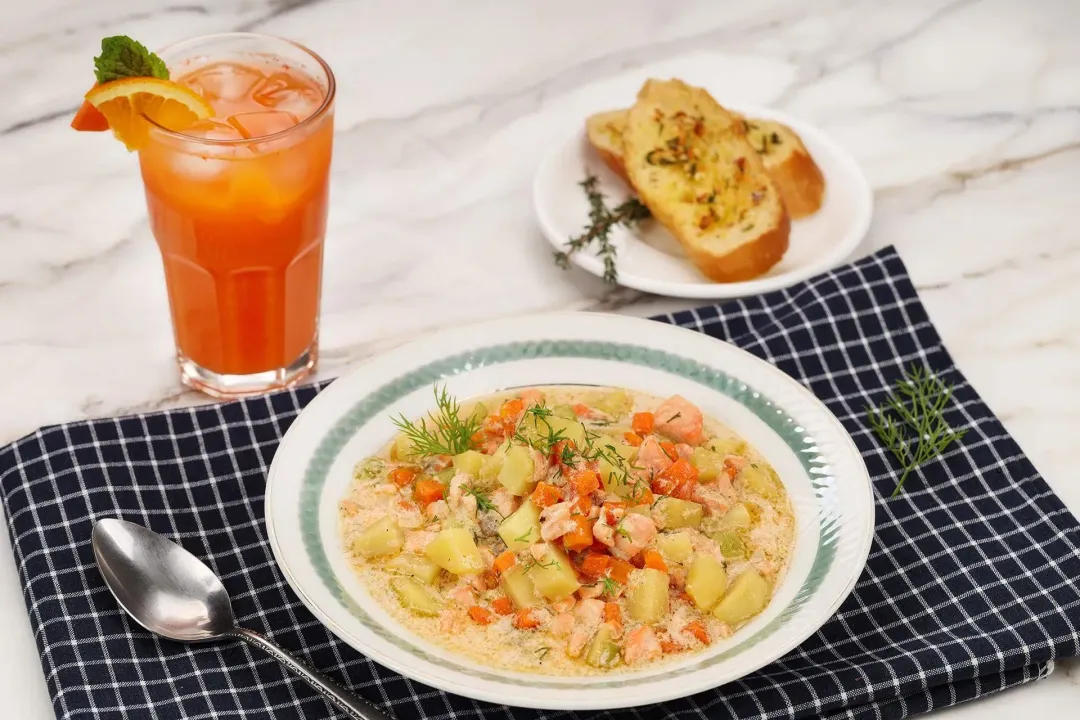 A deep plate containing salmon soup laid on a blue tablecloth near a spoon, a glass of orange carrot juice, and garlic toasted bread