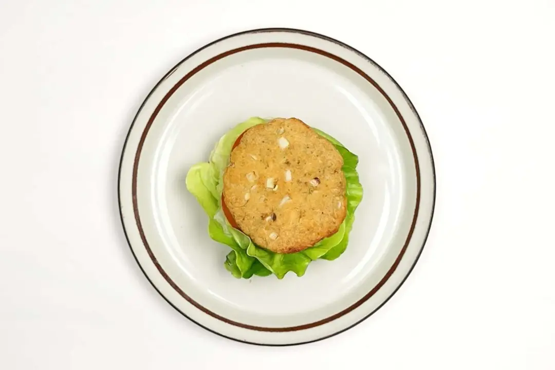A plate holding a hamburger without a top bun layering lettuce leaves, tomato slices, and a pale brown pattie on top of one another