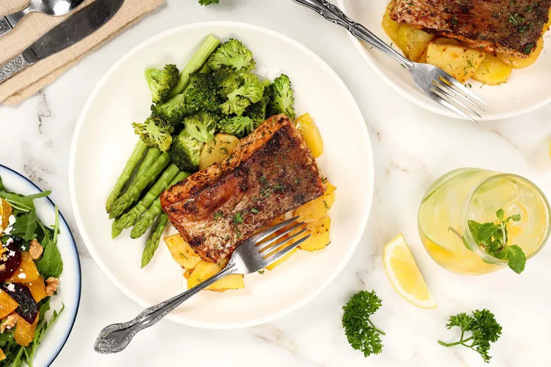 A high-angle shot of a plate of air fryer salmon, potato cubes, asparagus, and broccoli florets and a glass of pineapple juice