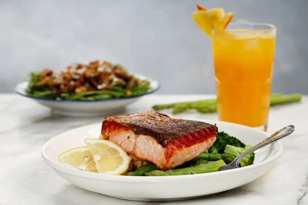 A plate of completed crispy-skin salmon with a plate of mushroom salad, a glass of orange pineapple juice and some fresh asparagus on the background