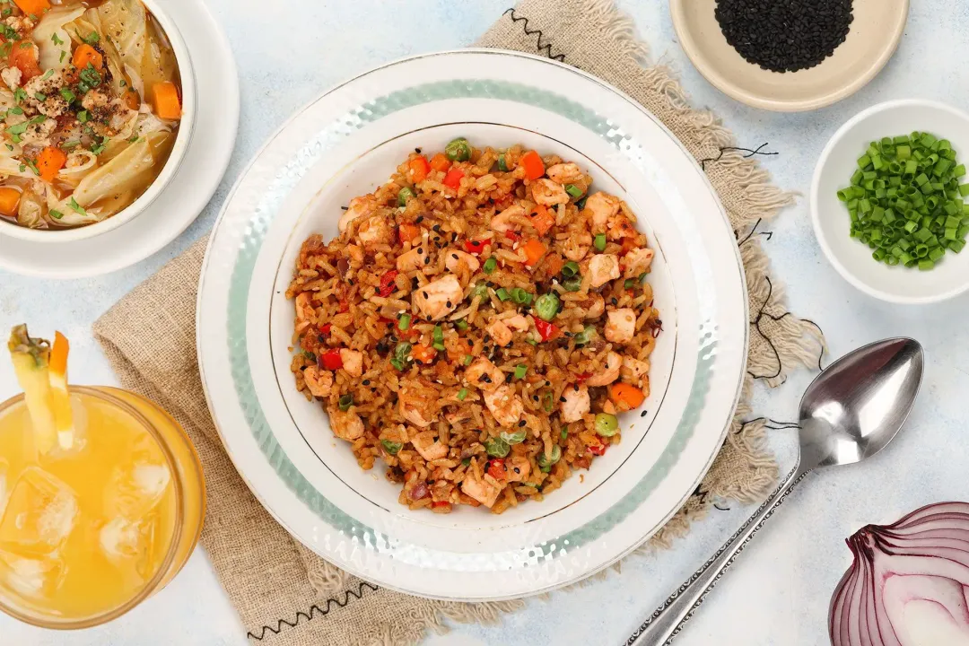 A plate of salmon and vegetable fried rice on a cloth, next to a soup, a juice, silverware, and herbs