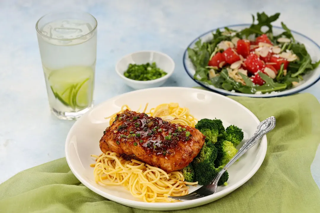 A plate of honey garlic salmon placed on a light green tablecloth with a palte of watermelon arugula salad and a glass of fresh lime juice on the background