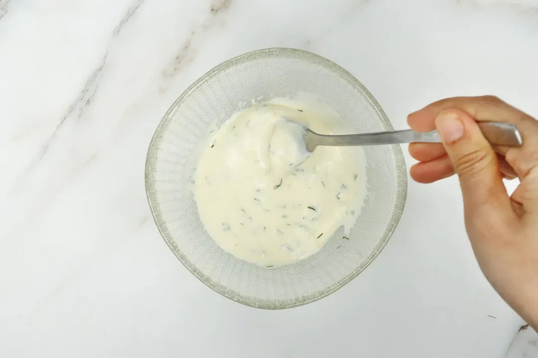 A hand whisking a creamy white sauce in a small glass bowl