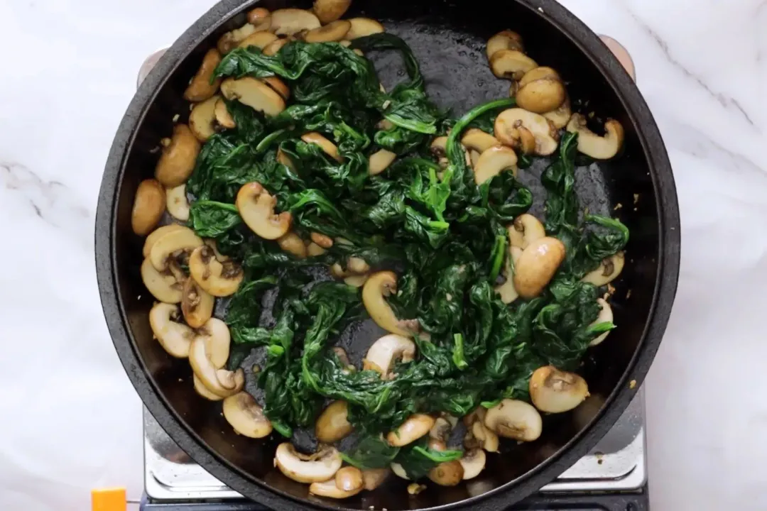 A skillet containing sliced cremini mushroom and cooked spinach leaves