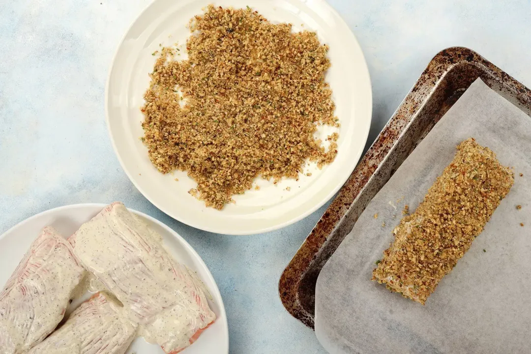 Glazed salmon filets are placed on one plate, a pecan crust mixture is placed on another plate, and a salmon filet that's dredged with the crust is placed on a baking sheet lined with parchment paper.