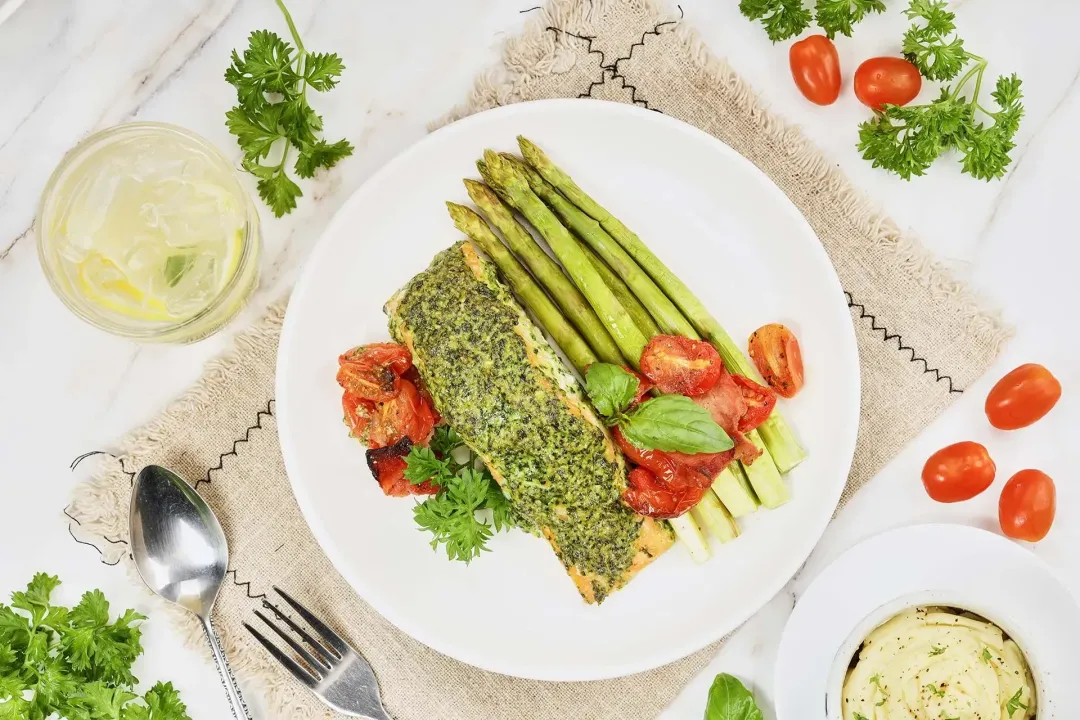 A plate of pesto salmon on a tablecloth surrounded by fresh parsley, a glass of lemonade, a bowl of mashed potatoes, fresh vegetables, and silver utensils