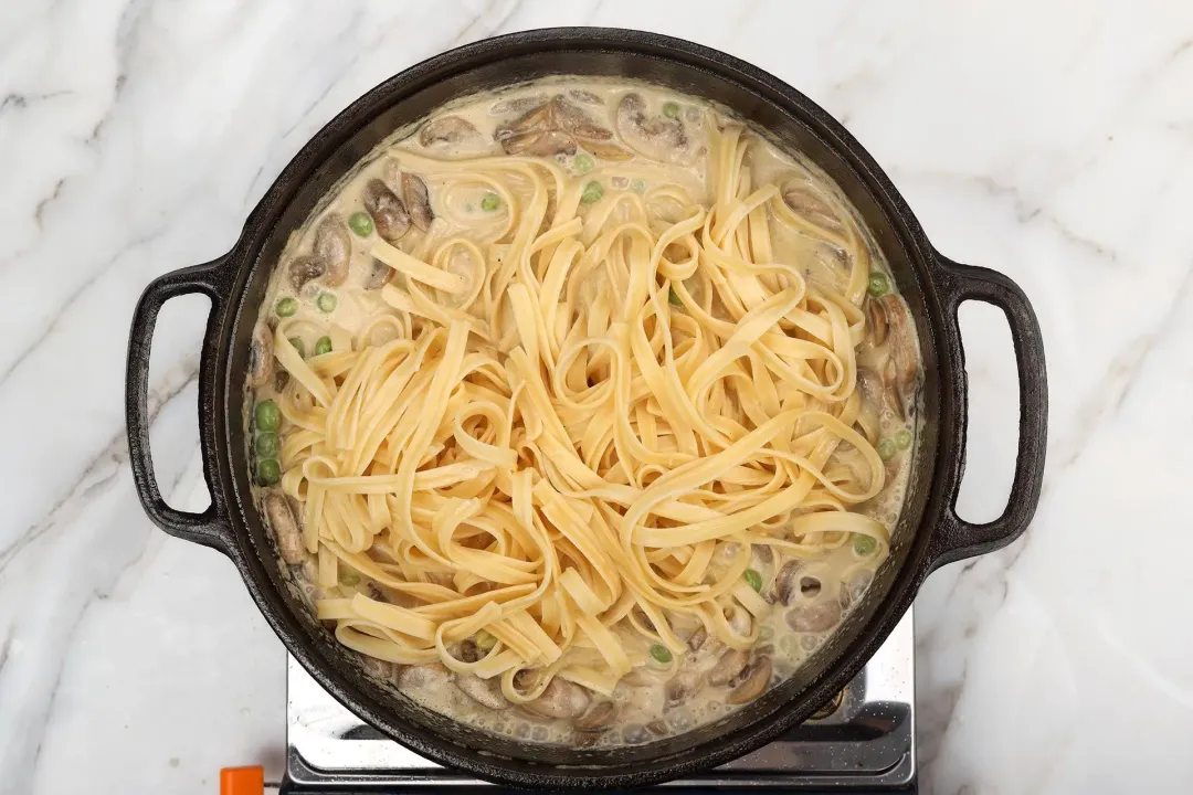 A skillet cooking fettuccine pasta in a mixture of heavy cream, mushrooms, and green peas