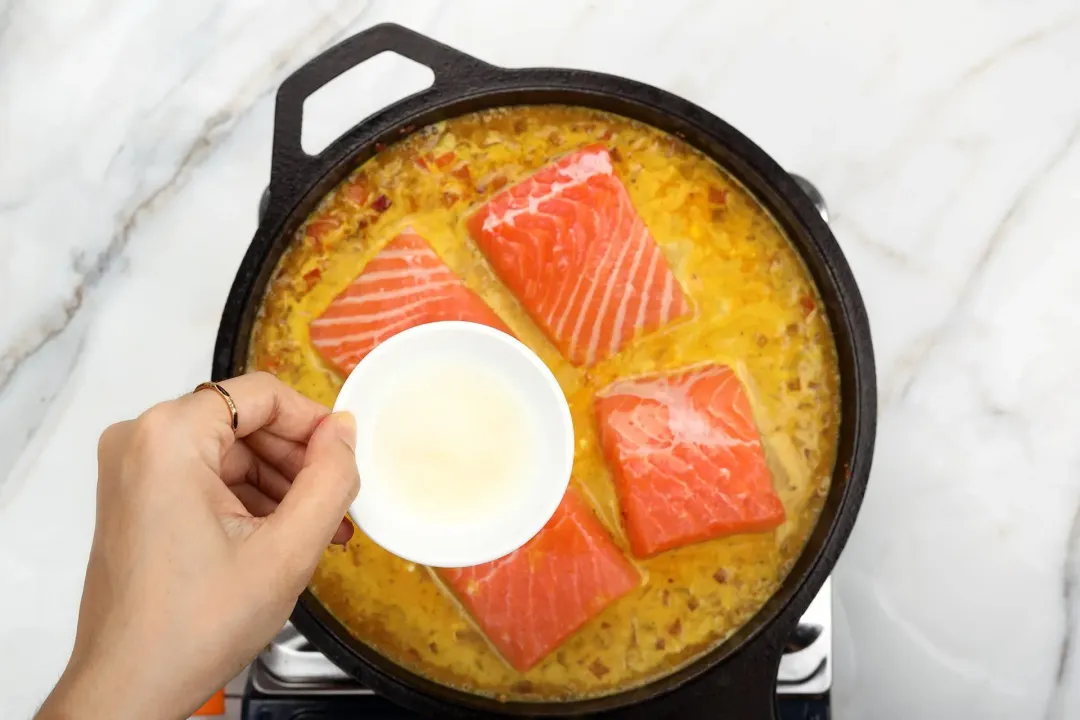 A hand holding a small dish containing lime juice over a skillet filled with four uncooked salmon fillets and curry