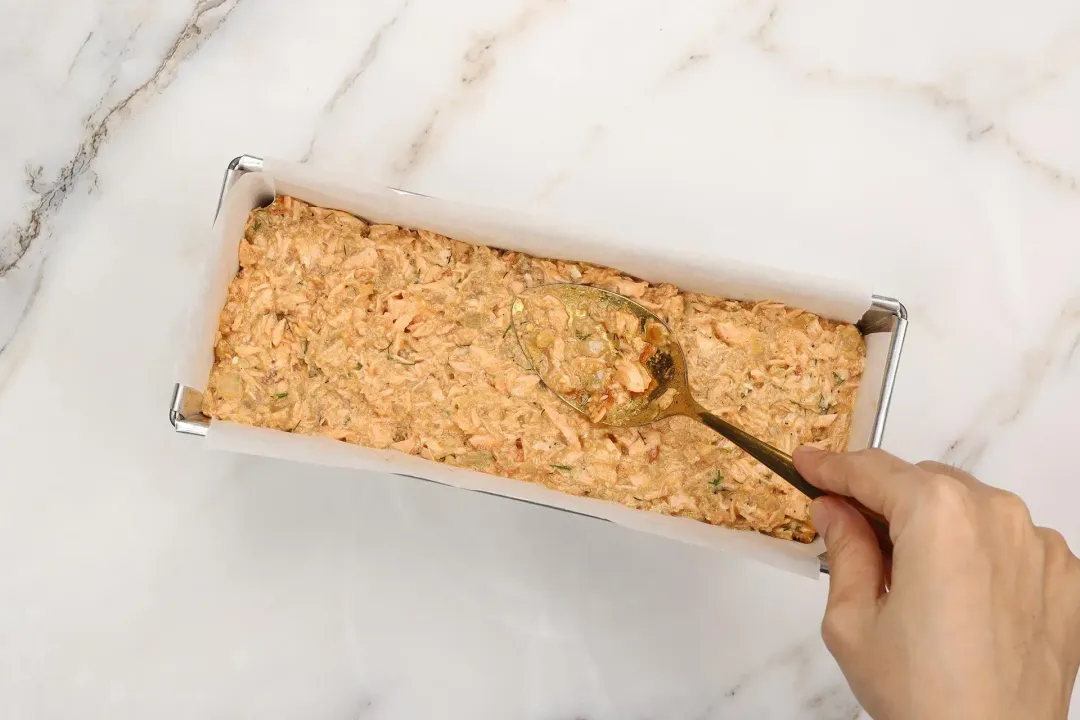 Salmon mixture spread into a rectangle mold lined with parchment paper by a gold spoon