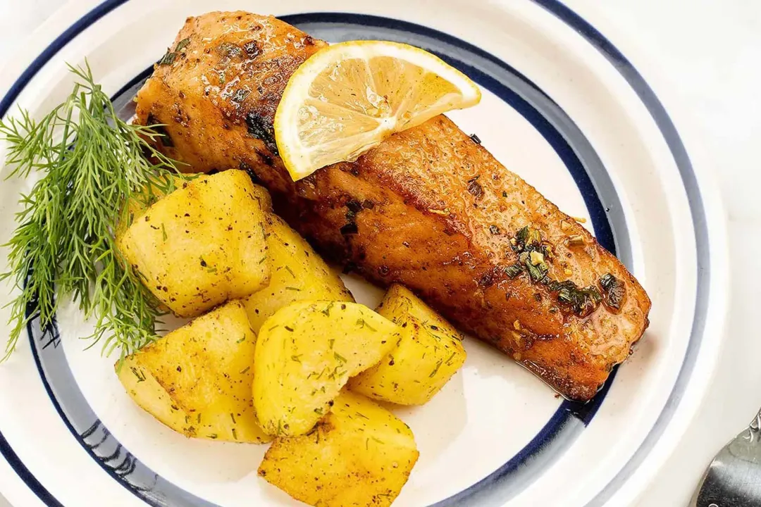 A white Chinese plate with a golden salmon filet topped with a lemon slice, potato wedges tossed with dill, and a dill sprig