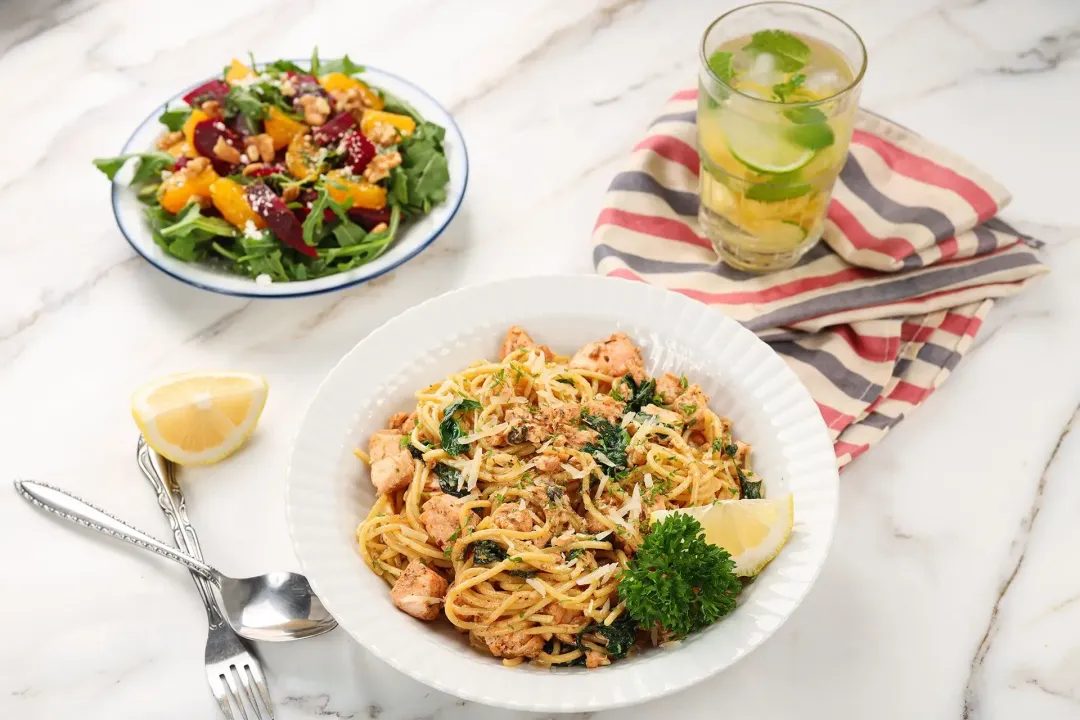 A plate of salmon pasta laid near a fork, a spoon, a lemon wedge, a beverage laid on a striped tablecloth, and a beet orange arugula salad