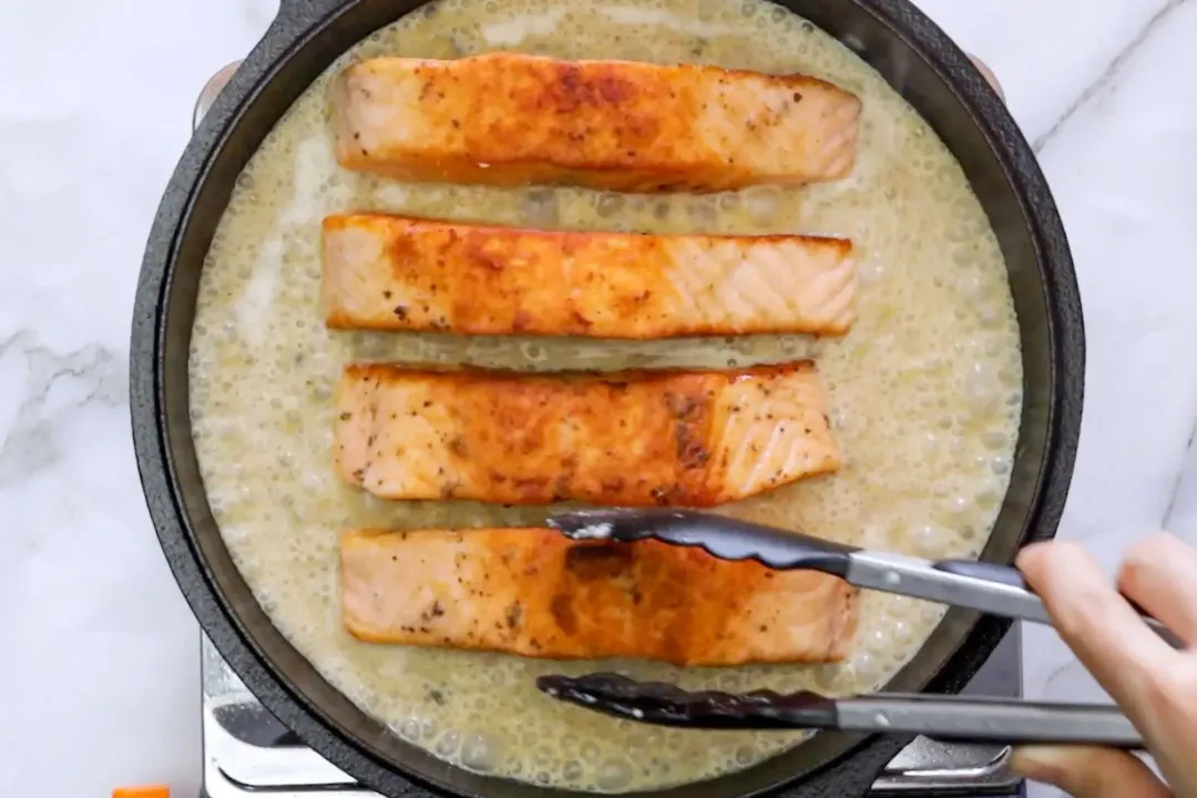 Four cooked salmon filets are being placed on a bed of a creamy mixture in a skillet, one of which is being held by a food grabber.