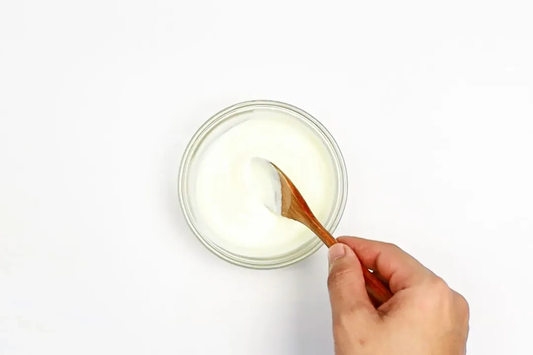 A picture taken from above of a jar of mayo-yogurt sauce with a hand stirring a spoon into the sauce.