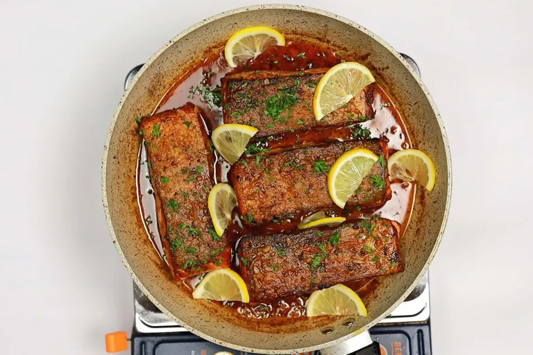A skillet on a stove with some oil, four golden-brown salmon fillets, lemon slices, and chopped parsley on top.