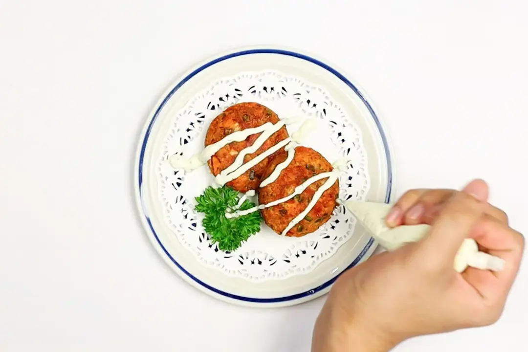 A hand is drizzling the white mayo-yogurt sauce over the two fried salmon patties in a plate garnished with parsley.
