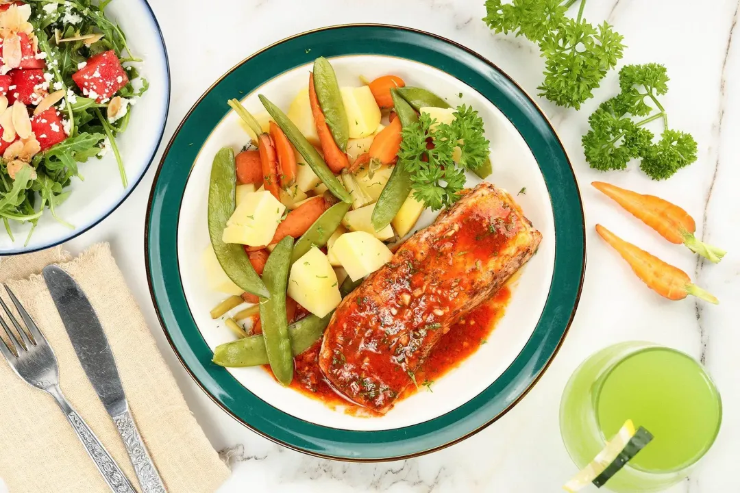 A plate of Instant Pot salmon surrounded by a glass of juice, a plate of salad, some fresh vegetables, and dining utensils