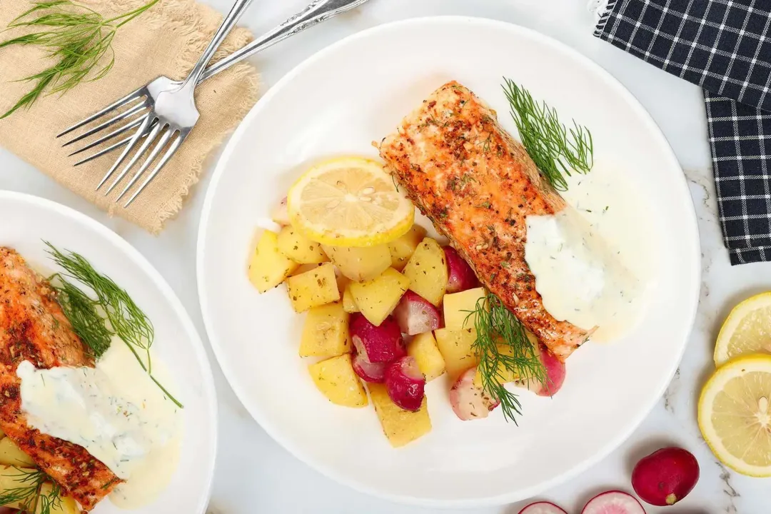 A high-angle shot of a plate of cooked salmon fillet, potato cubes, lemon slice, radish cubes, fresh dill, and a creamy white sauce