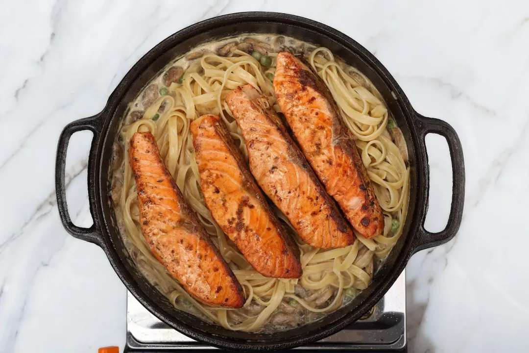 A skillet cooking four salmon fillets with fettuccine pasta