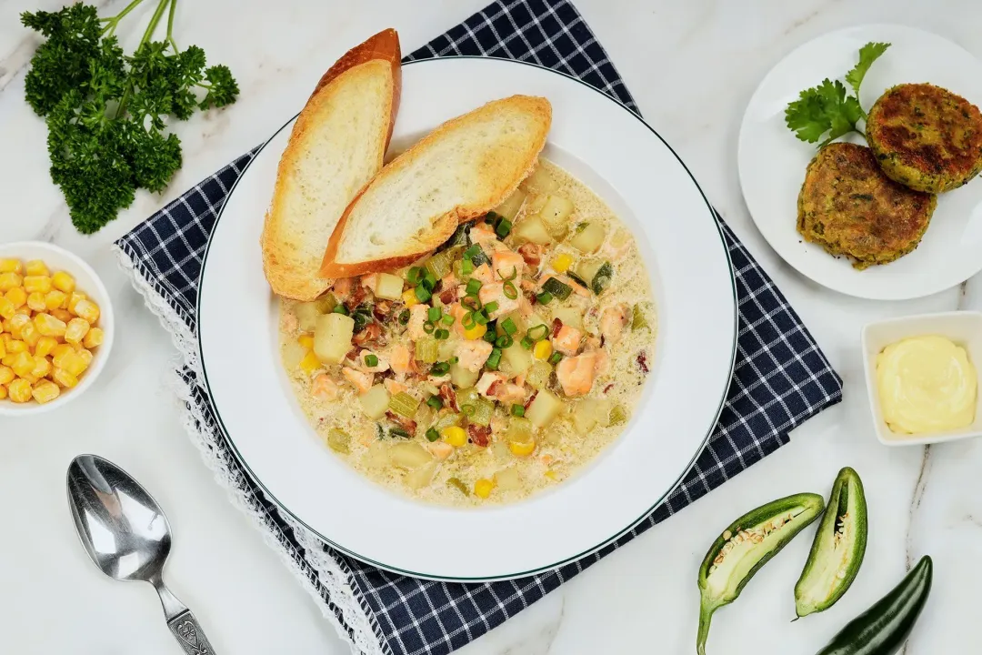 A plate of salmon chowder on a blue checkered tablecloth surrounded by falafel, halved jalapeno, parsley and some corn kernels