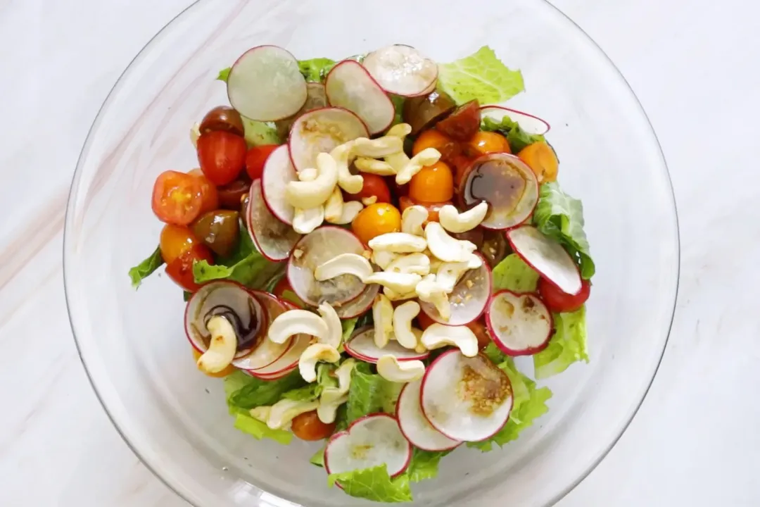A glass bowl of colorful salad with lettuce, radish slices, tomato halves, and cashews.