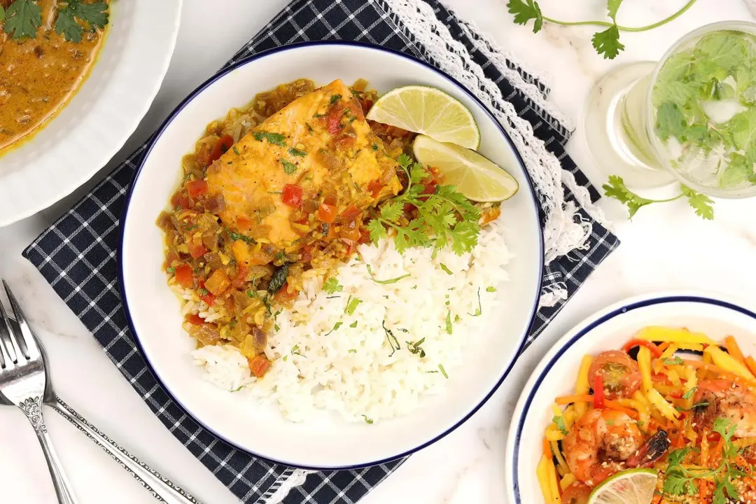 A flatlay of a dish containing salmon curry, white rice, and lime wedges laid next to Thai salad and lime juice