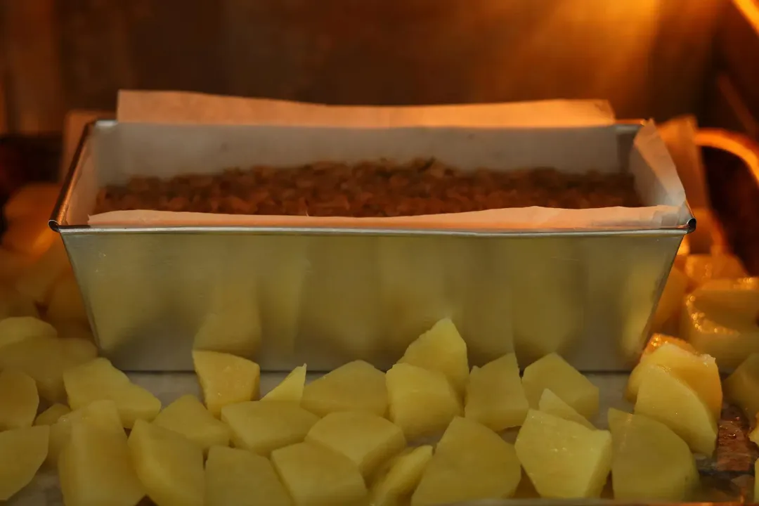 A rectangle aluminium mold containing salmon mixture baking in the oven along with large potato cubes