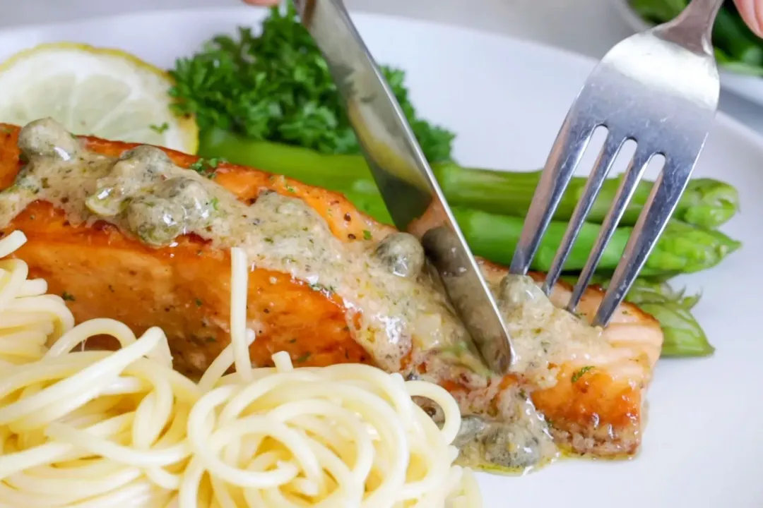A salmon filet is being held by a fork and cut by a knife, on top of which is a creamy sauce with some capers, and on the side there are a cooked pasta, cooked asparagus, a lemon slice, and parsley.