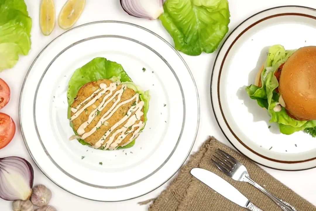 A flatlay of two plates holding hamburgers, a fork, a dinner knife, lettuce leaves, red onion, and tomato slices