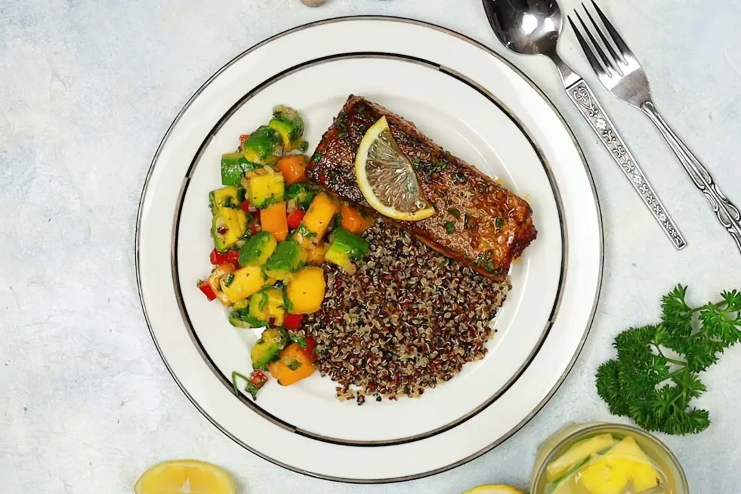 A picture of a plate of pan-seared salmon fillet, quinoa, and mango-avocado salsa next to a spoon, a fork, lemon slices, parsley, and a glass of juice.