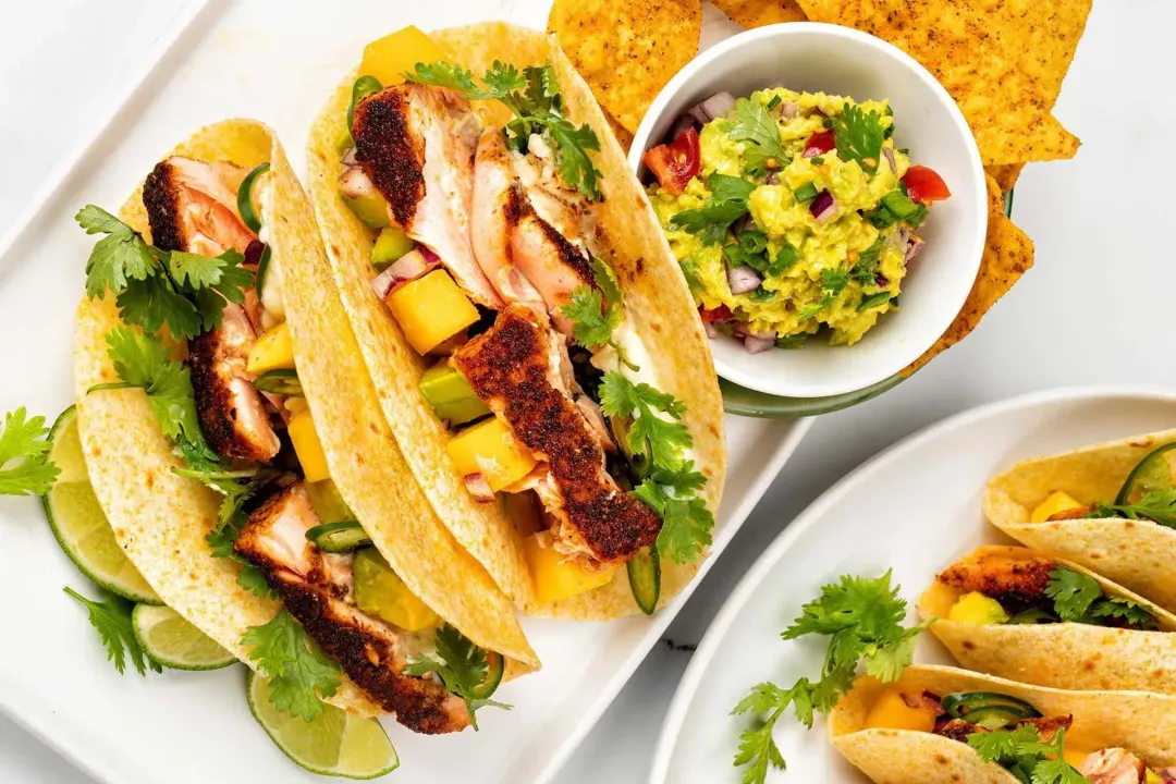 A square plate and a round plate with some blackened salmon tacos, salad, tortilla chips, lime slices, and fresh parsley.