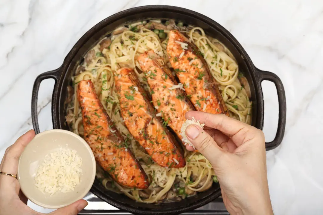 A hand sprinkling some shredded parmesan cheese into a skillet that contains cooked salmon fillets and fettuccine pasta