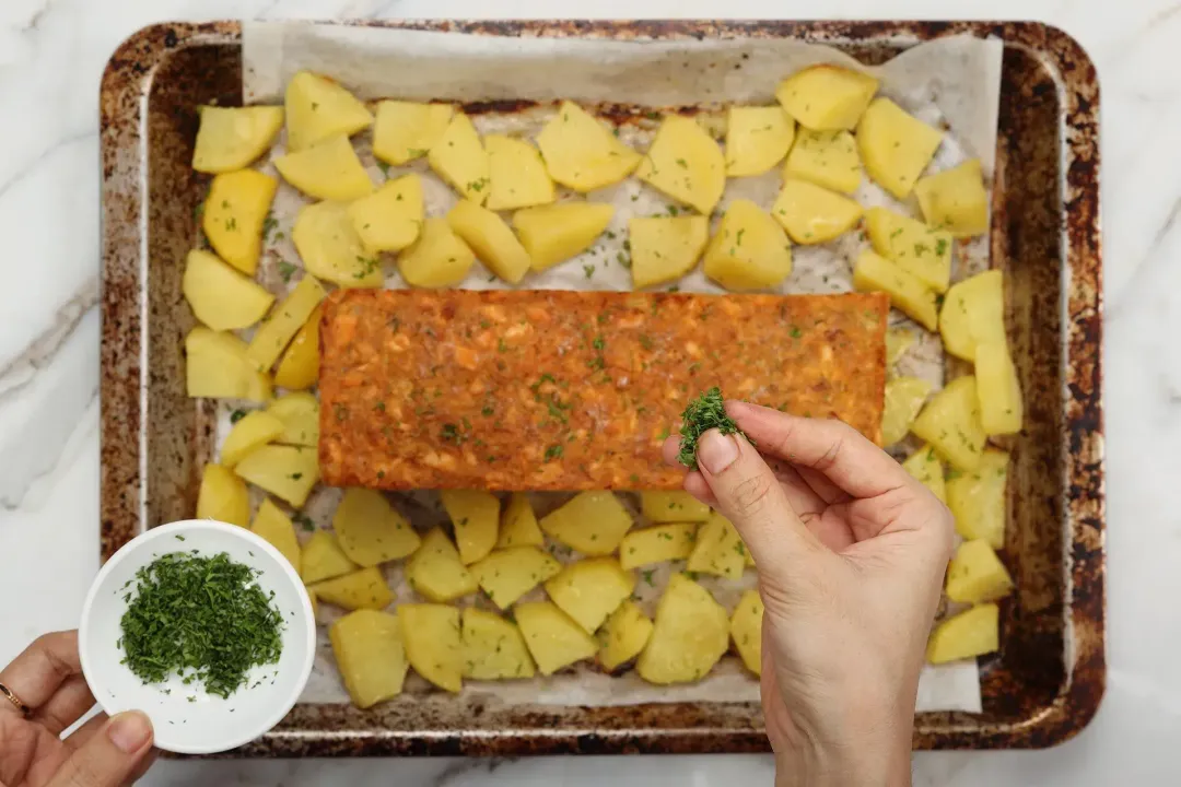 A hand preparing to sprinkle chopped herb onto a salmon cake laid in the center of a baking sheet lined with parchment paper and surrounded by potato cubes