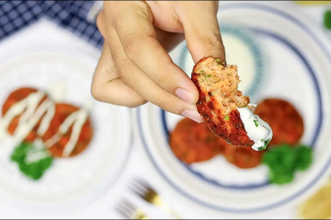 A close up picture of a hand holding out a piece of fried salmon patty dipped in mayo-yogurt sauce in the background of two plates of salmon patties.