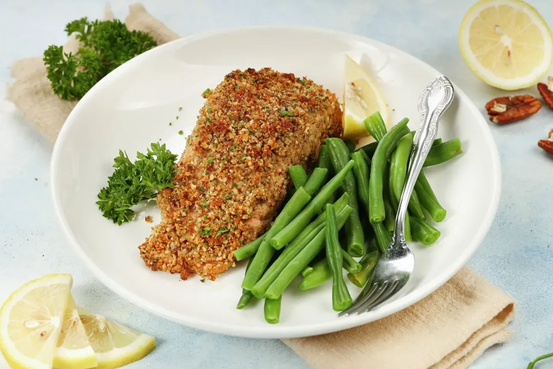 A filet of pecan-crusted salmon on a serving plate, with cooked green beans, a few lemon slices, parsley, and a fork on the side.