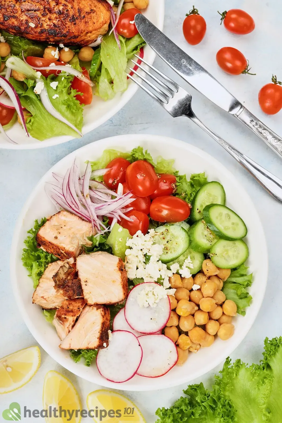 A round white plate containing a salmon salad laid near lemon wedges, cherry tomatoes, a dinner knife, a fork, and another plate of salmon salad