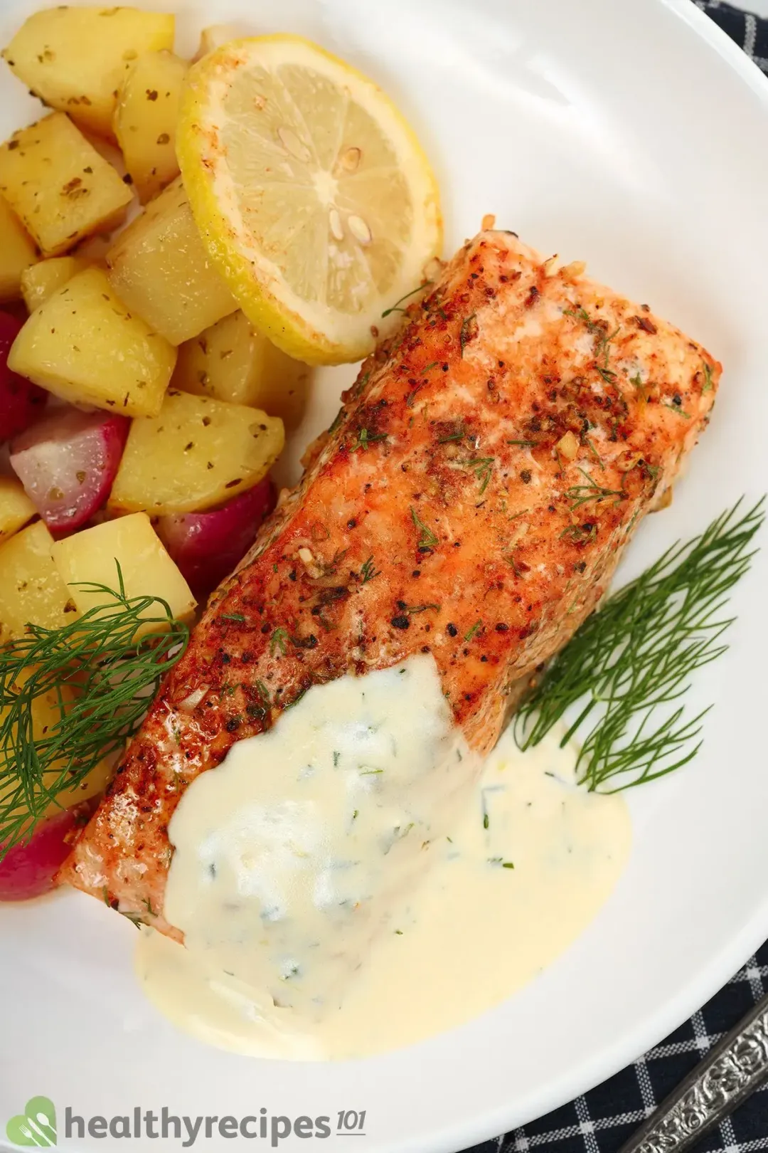 A cooked salmon fillet is partially covered in a creamy white sauce laid near fresh dill, potato cubes, and a lemon slice