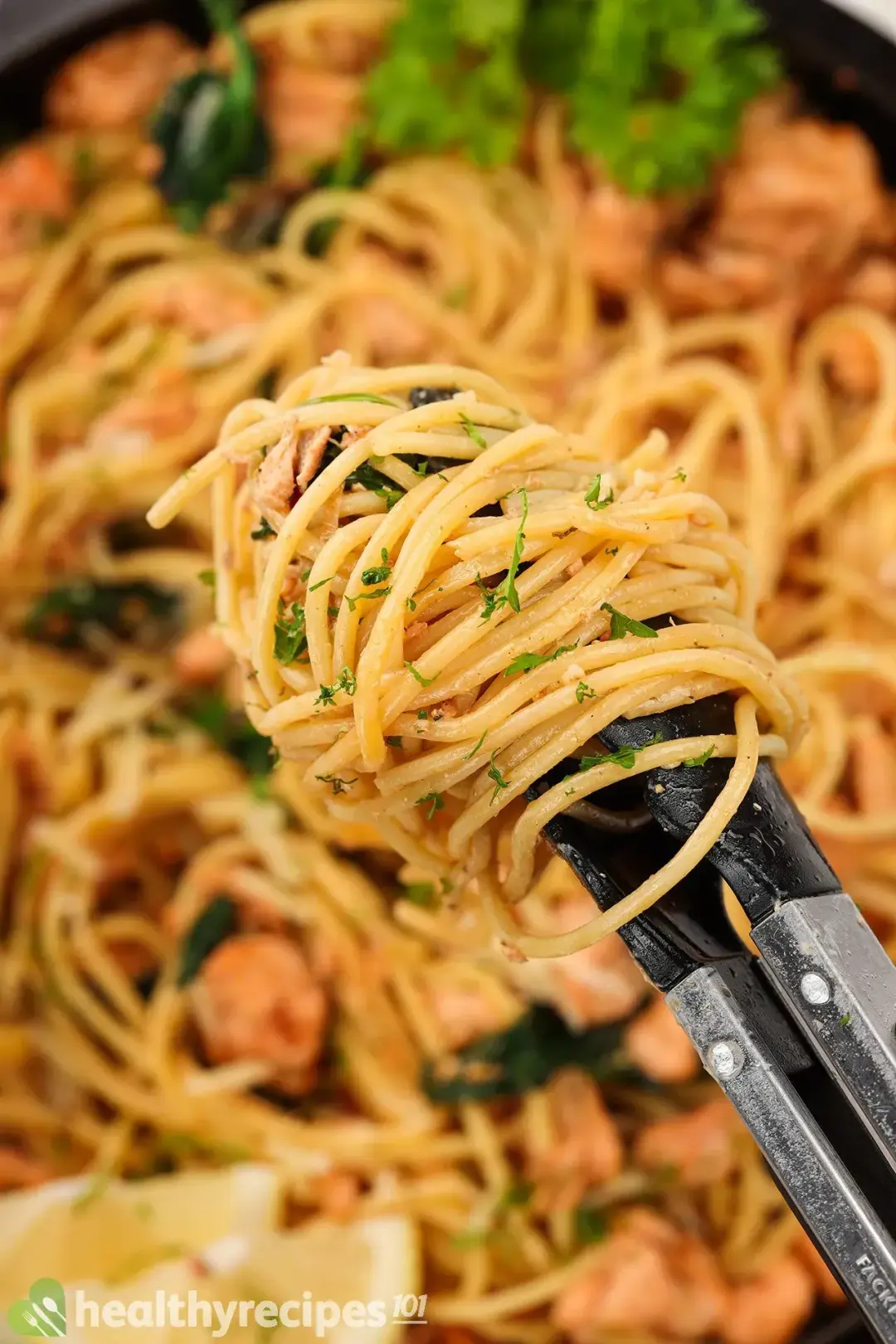 A pair of tongs with spaghetti and chopped parsley wrapped around it with a skillet containing spaghetti salmon in the blurred background