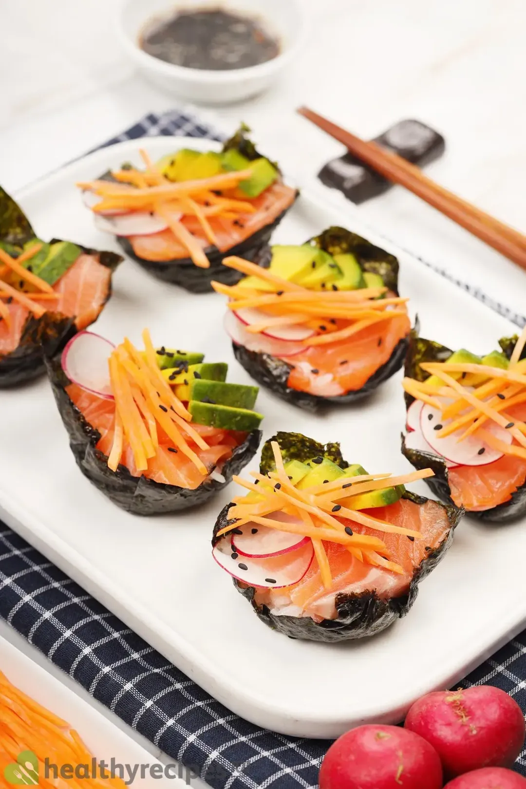 Six sushi cupcakes lined neatly on a white plate, above a blue checkered cloth and next to red radishes