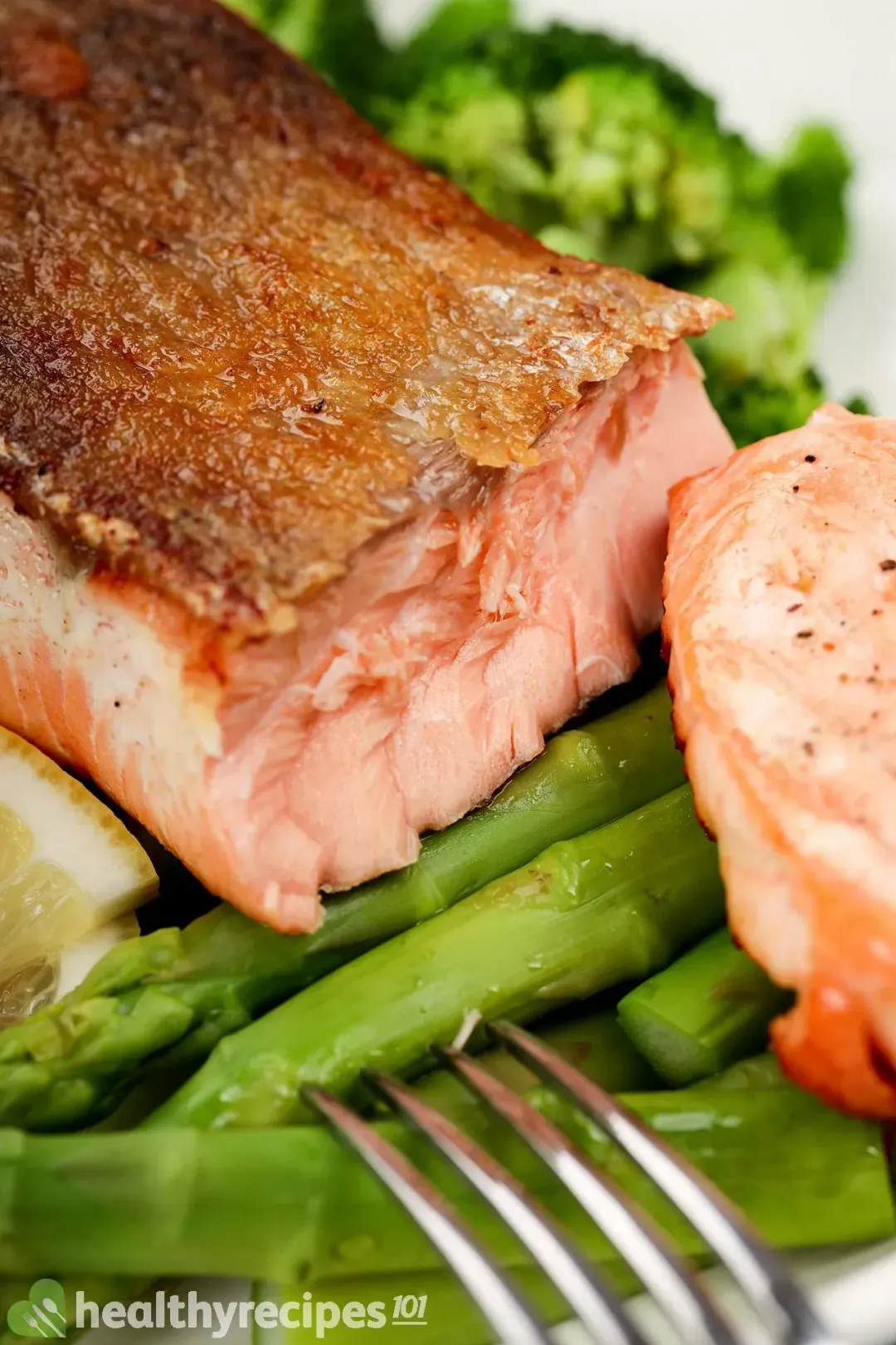 A close-up shot of crispy-skin salmon on a bed of boiled asparagus and broccoli with two slice of lemon on the side