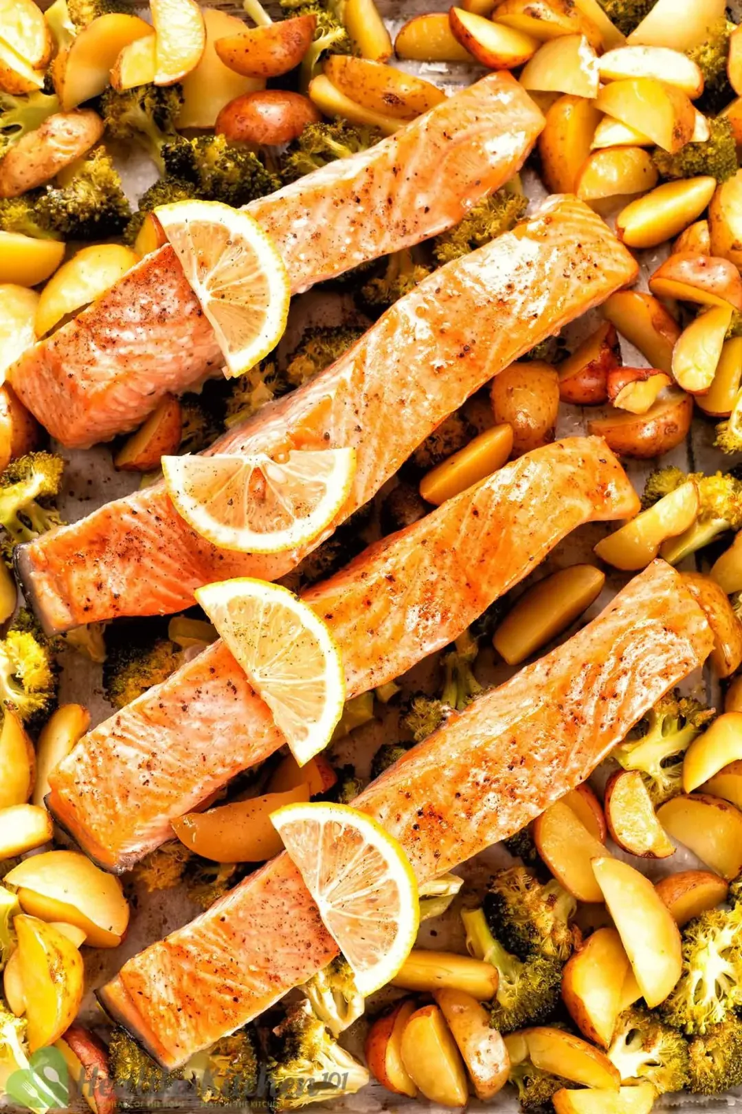 Four marinated and oil-coated salmon filets are assembled on a baking sheet, topped with four lemon slices and surrounded with potato wedges and broccoli pieces.