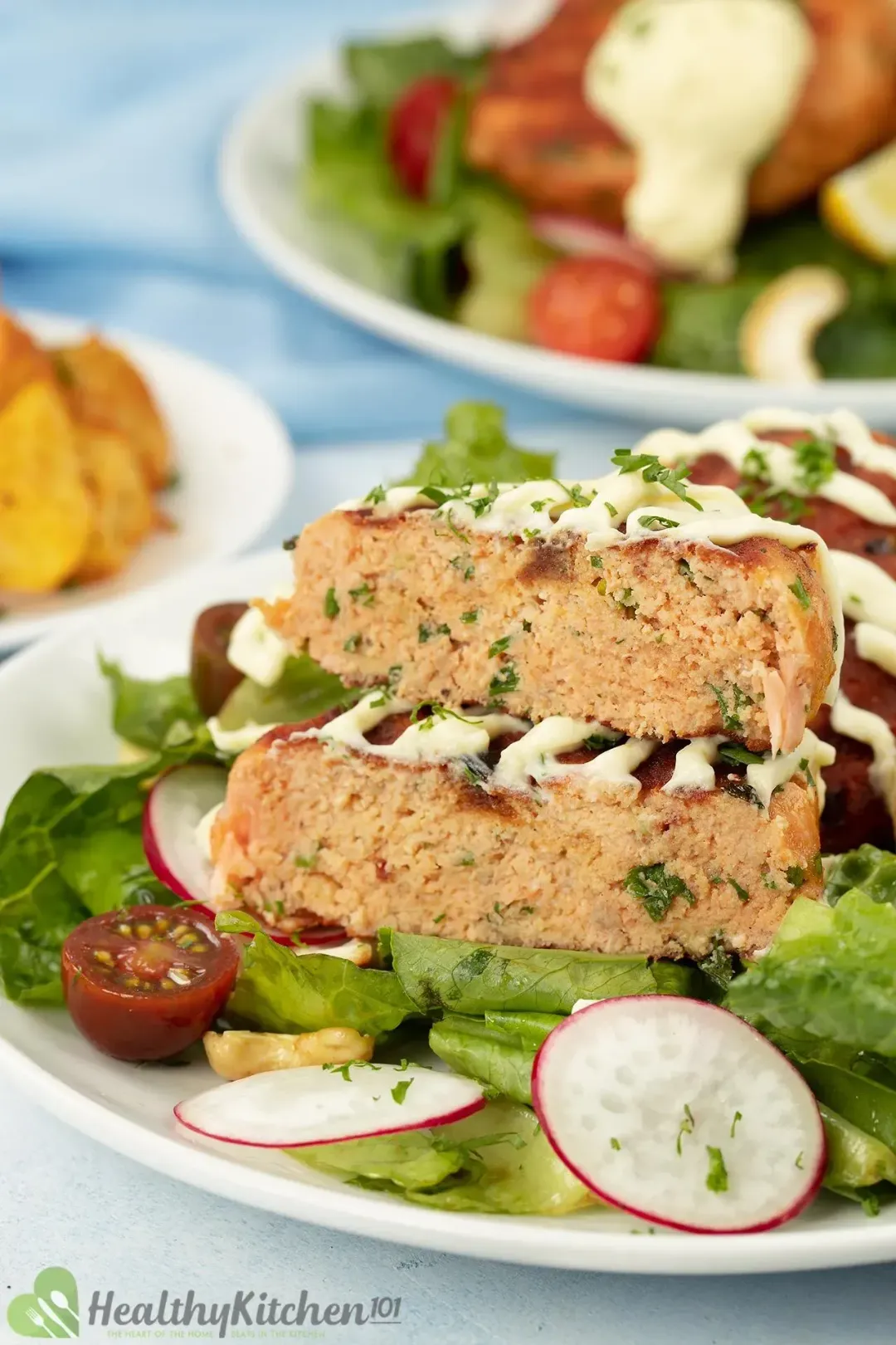 Pieces of salmon croquettes drizzled with the white sauce on a plate with fresh veggies and greens.