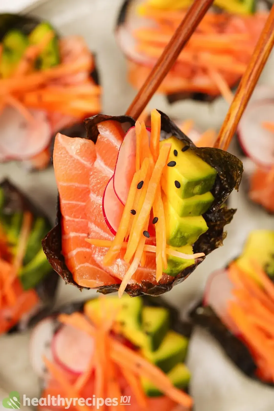 A pair of chopsticks holding a sushi cupcake of salmon, carrot, avocado, and radish hovering six other sushi cupcakes