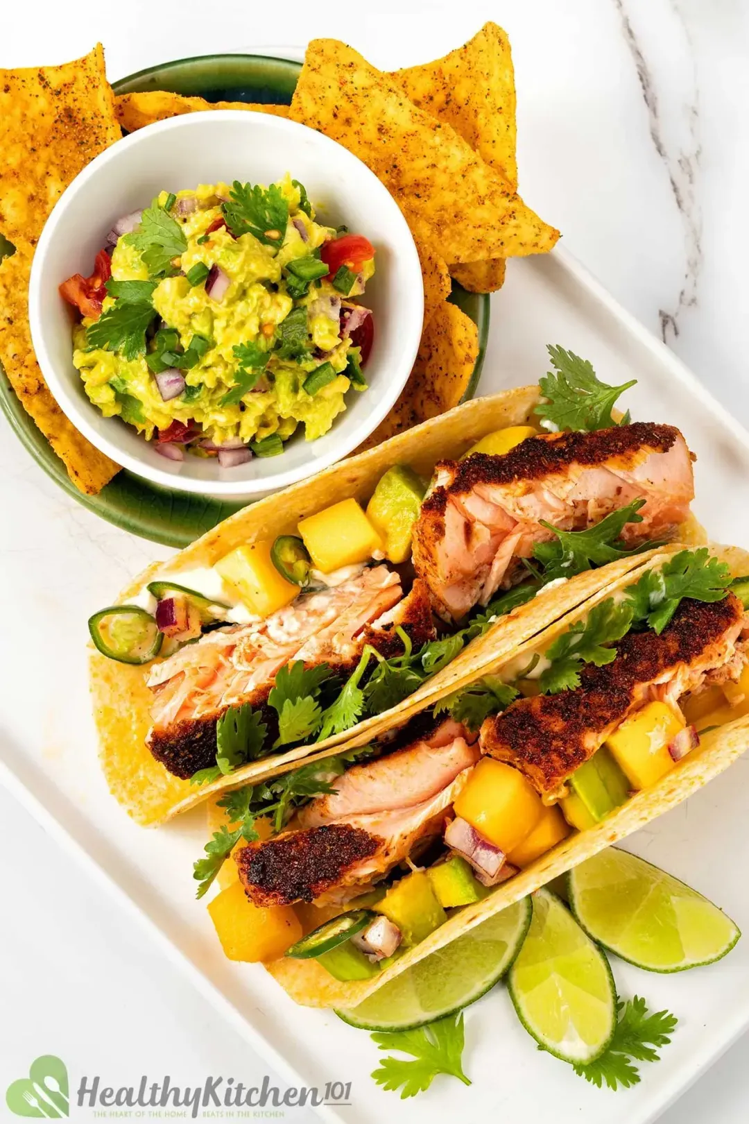 A picture of two blackened salmon tacos on a white plate decorated with lime slices and parsley on the right side of a small bowl of avocado mango salsa and some tortilla chips.
