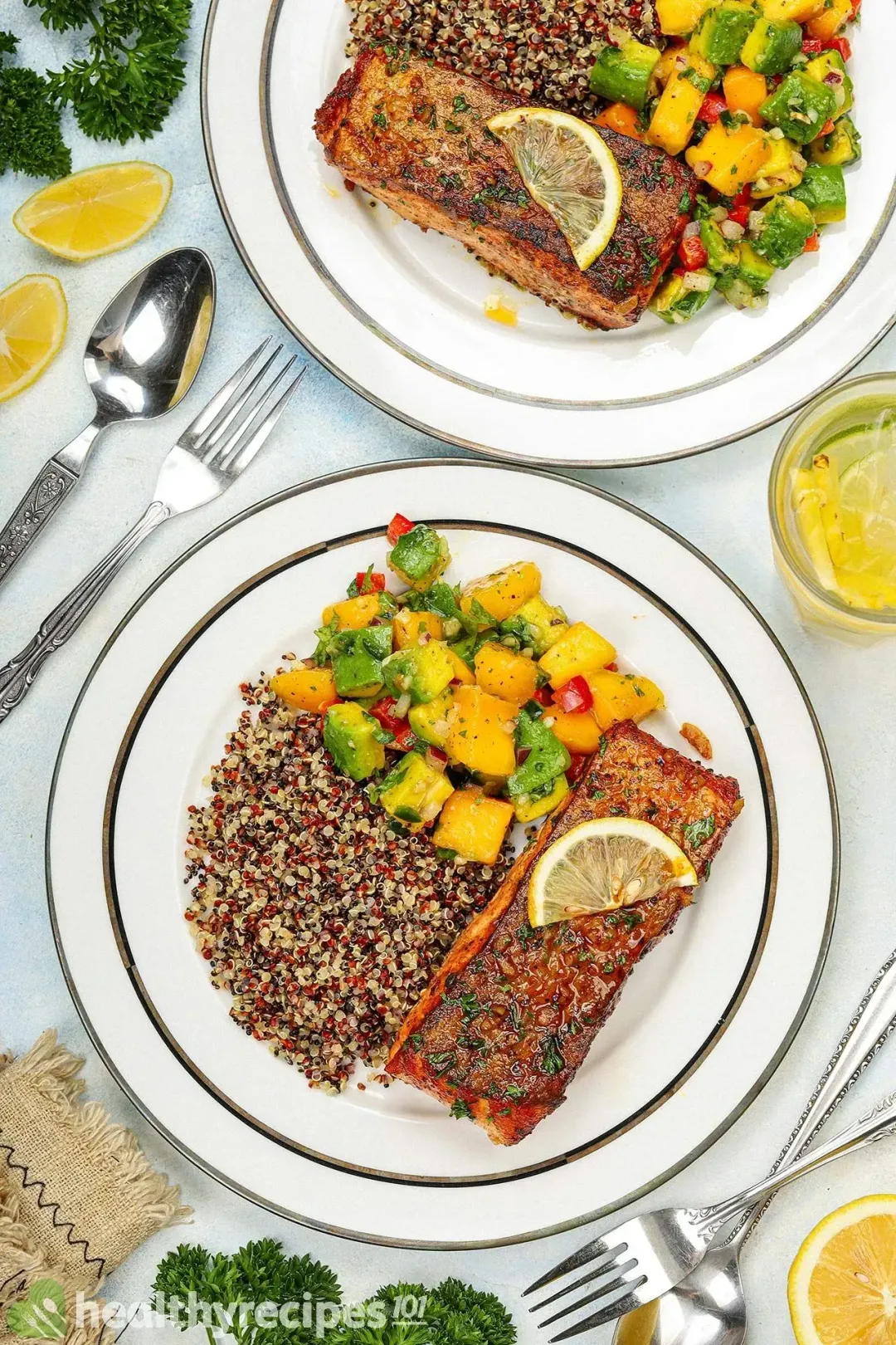 The picture of a plate of pan-seared salmon fillet, quinoa, and mango-avocado salsa next to another similar plate, some spoons, forks, lemon slices, herbs, and a glass of juice.