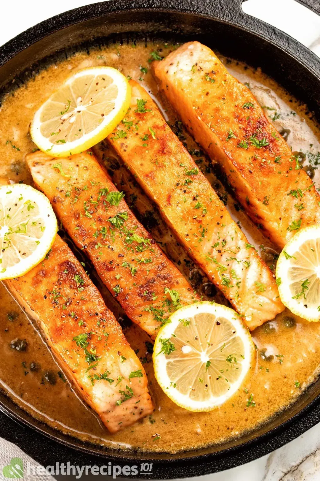 Four salmon filets sitting in a creamy cooking juice in a skillet are topped with four lemon slices.