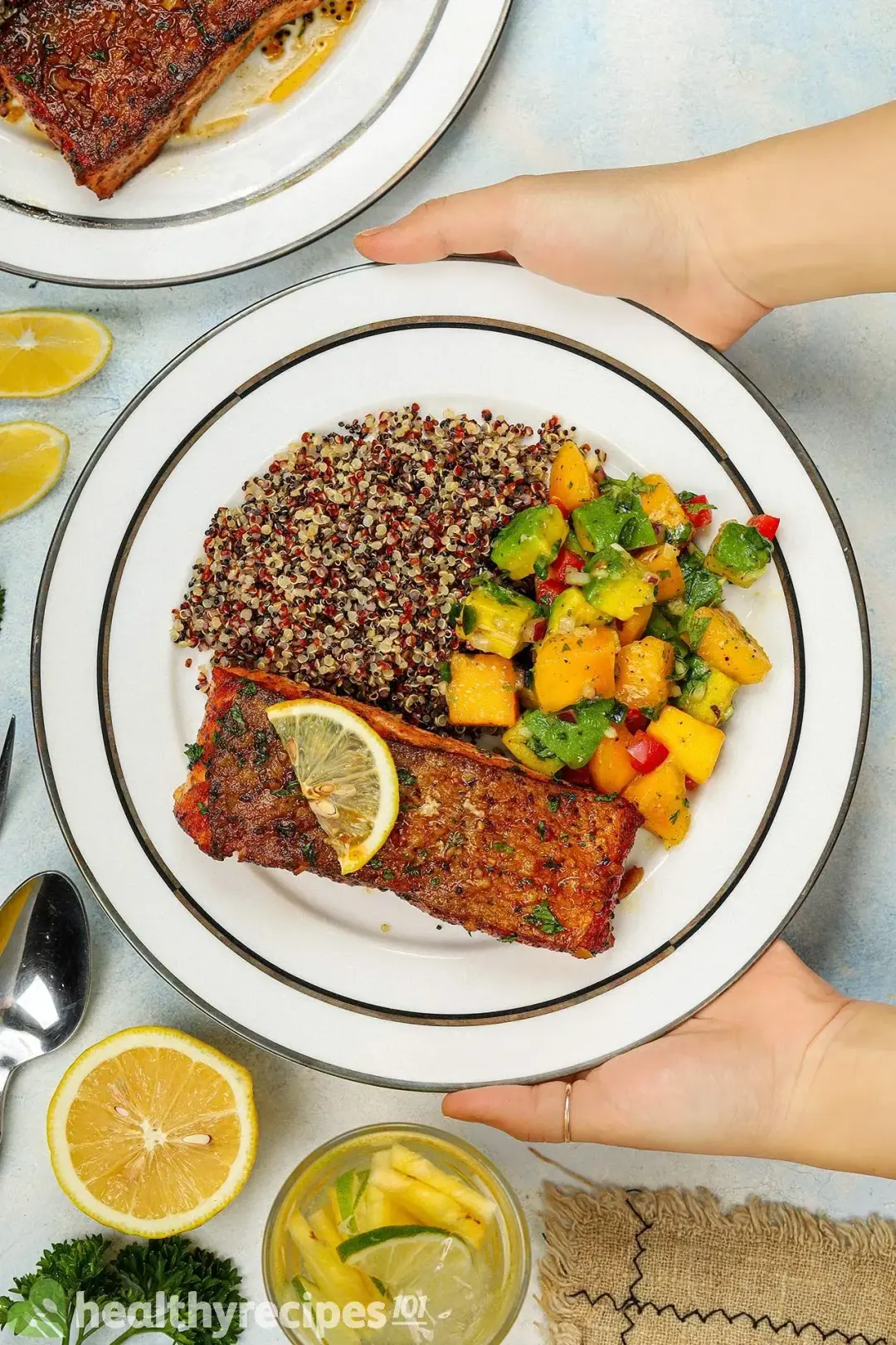 Pan-seared salmon fillet, mango-avocado salsa, and mixed quinoa in a plate held out by a hand, next to a cup of lime and pineapple juice, half a lemon, and a spoon.