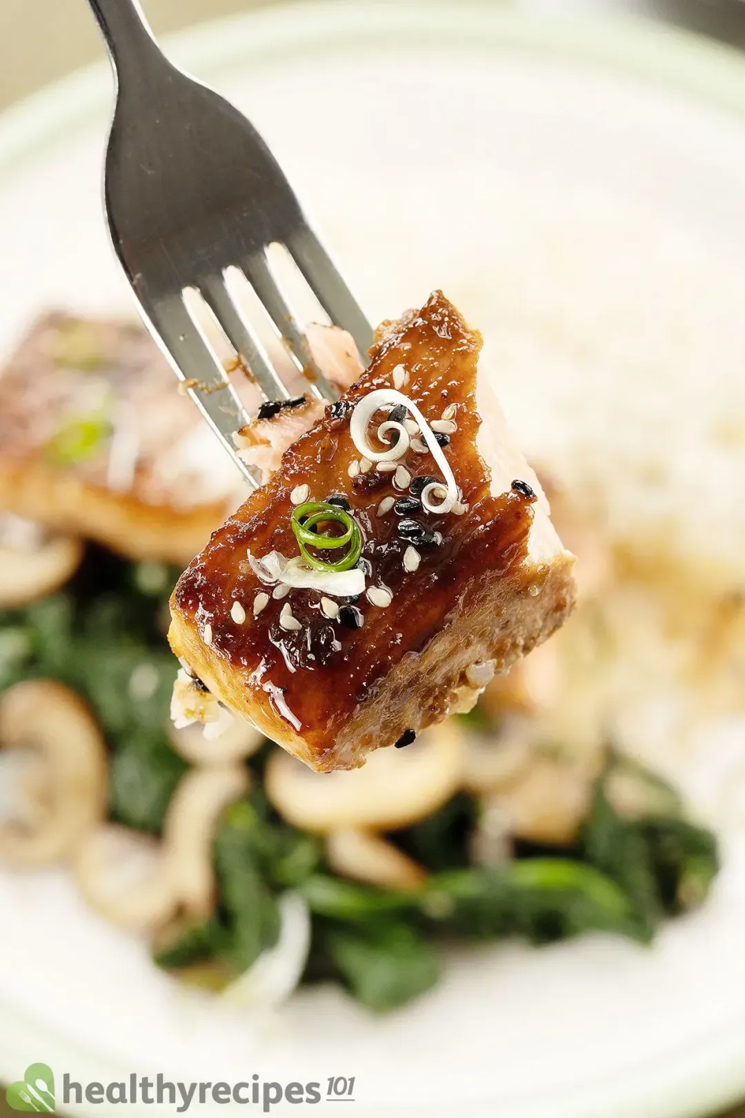 A fork lifting a piece of salmon covered in a glossy brown miso sauce, a piece of chopped scallions, and some sesame seeds