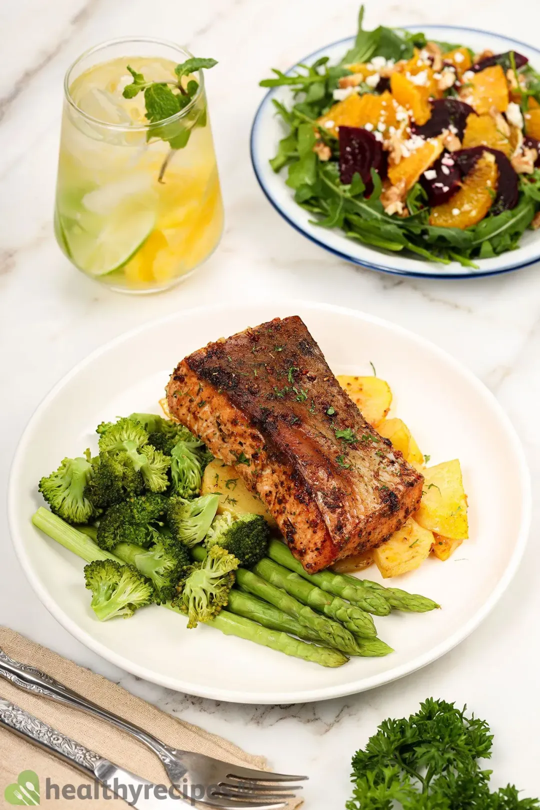 A plate of air fryer salmon, potato, broccoli florets, and asparagus laid near a plate of salad and a glass of pineapple water