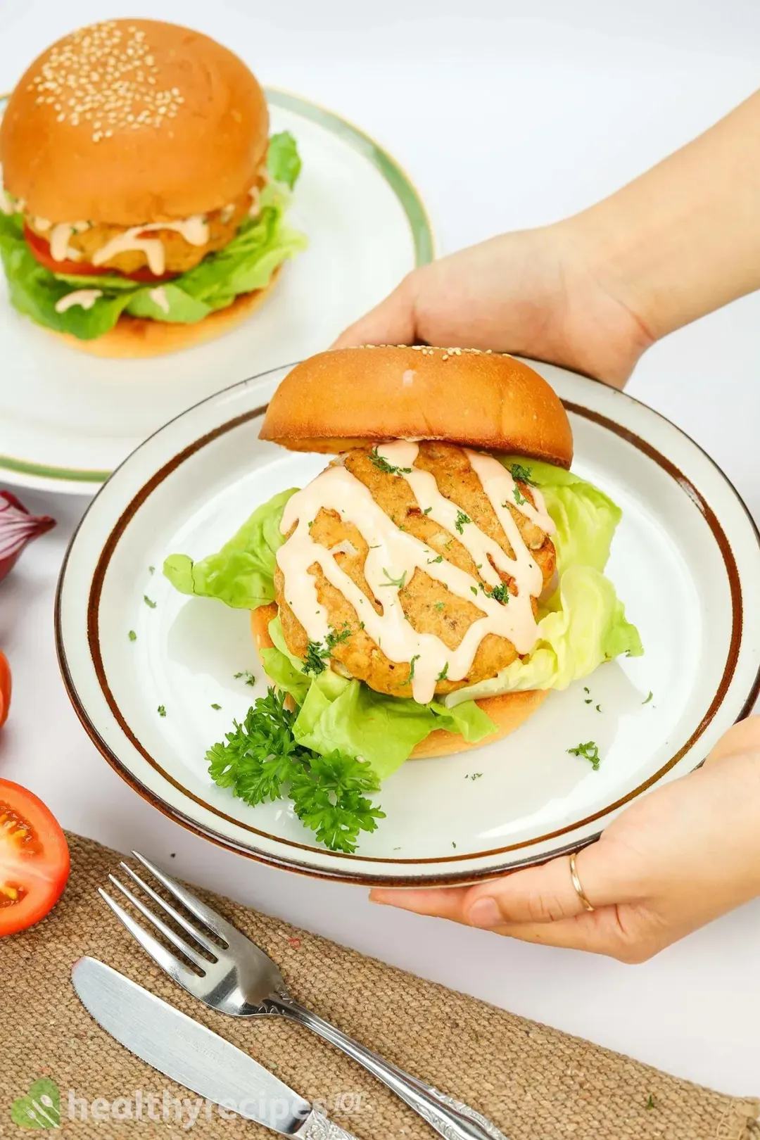 Two hands holding a plate containing a hamburger with the top bun fallen on the side, revealing a pattie covered in a sauce and chopped herbs