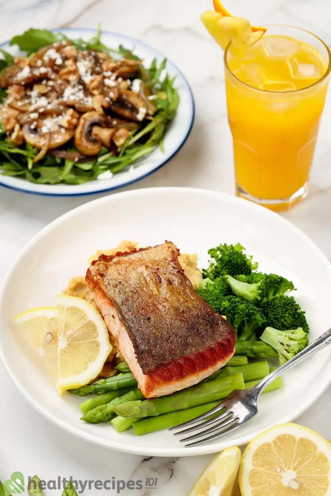 A plate of crispy-skin salmon served with a plate of mushroom salad and a glass of orange pineapple juice